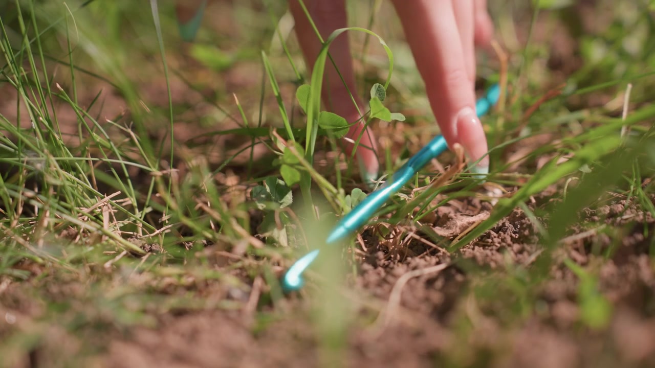 Close up of hand using blue crochet hooks to work on yarn in grass demonstrating crafting techniques with soft natural light and detailed focus on tool and material interaction. scene