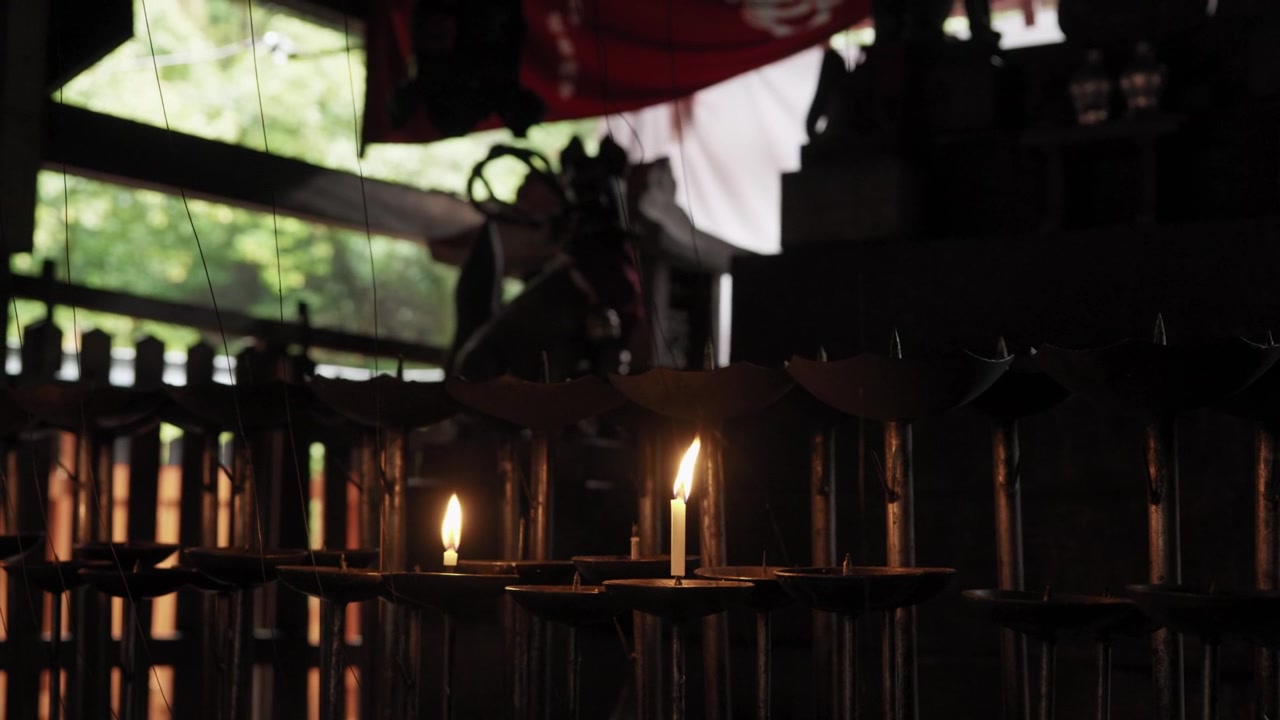 Two lit candles glowing on holders in a dark Japanese shrine interior with green foliage seen beyond in Japan