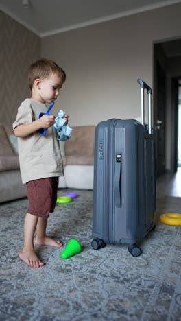 lovely kid holding a shirt comes up to a big suitcase standing in the middle of the room. toddler puts on sunglasses which he picks from the floor. vertical video.