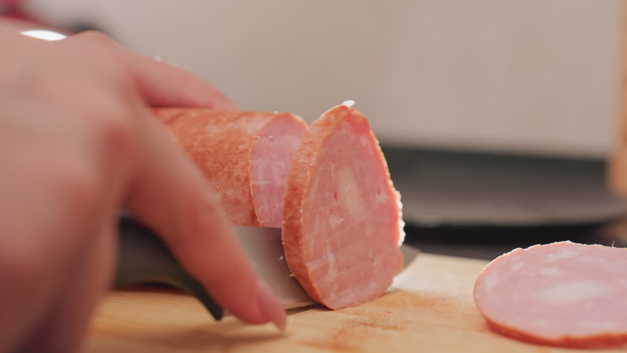 close up hands slicing pink sausage on wooden board using sharp kitchen knife in soft natural lighting with blurred background during food preparation in kitchen showing fresh meat