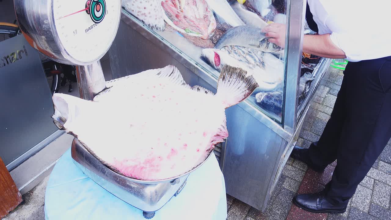 A bustling fish market with fresh seafood on display and a large flatfish on a scale.