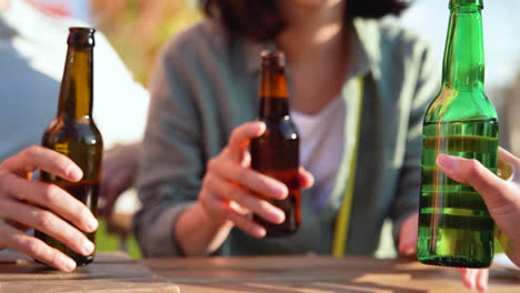 close up of three unrecognizable friends toasting with beer while sitting at table outdoors in a sunny day