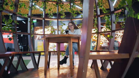 young lady seated in decorative sit-out, focused on her phone with pink and mint green shopping bags by her side, glowing lights in background, while someone passes behind in a vibrant atmosphere