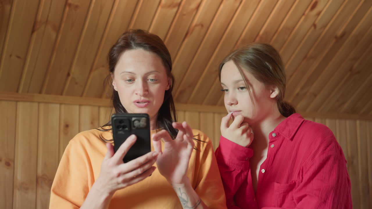 two girls sit closely in wooden room, one in orange shirt operating smartphone with focus while other in red shirt looks at screen pensively, both appearing calm and engaged in digital content