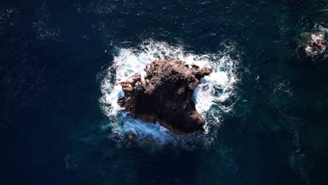 aerial top down view of waves smashing a cliff in middle of blue ocean