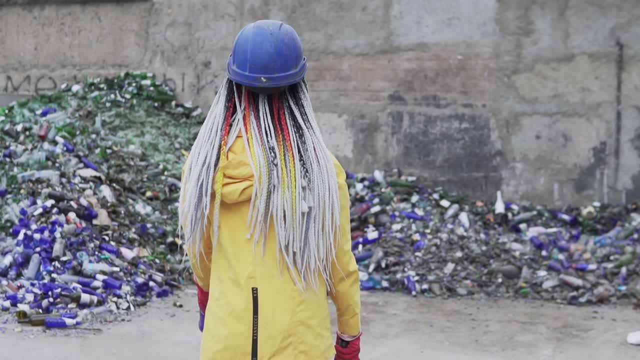 mujer joven con sombrero duro de pie contra la pila de vidrio roto, botellas usadas junto a la pared. niña con chaqueta amarilla rompiendo viejas botellas de vidrio para su reciclaje. vista rara. cámara lenta