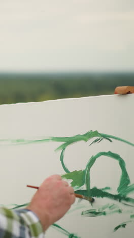se ve a un pintor, vestido con una camisa a cuadros, aplicando suavemente pintura verde a un gran lienzo blanco en un entorno al aire libre, centrándose en los delicados trazos de verde en el lienzo