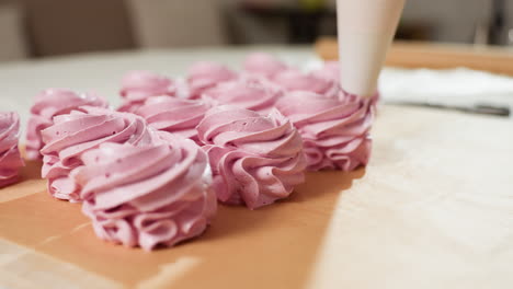 close up of cupcake preparation process showing piping bag squeezing swirls of pink dough onto brown baking paper, cupcakes aligned in neat row ready for baking, soft focus background