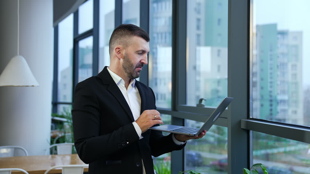 Male entrepreneur standing at the window and holding laptop in front of him. Business talk on the computer. Man talking and gesturing in front of notebook.
