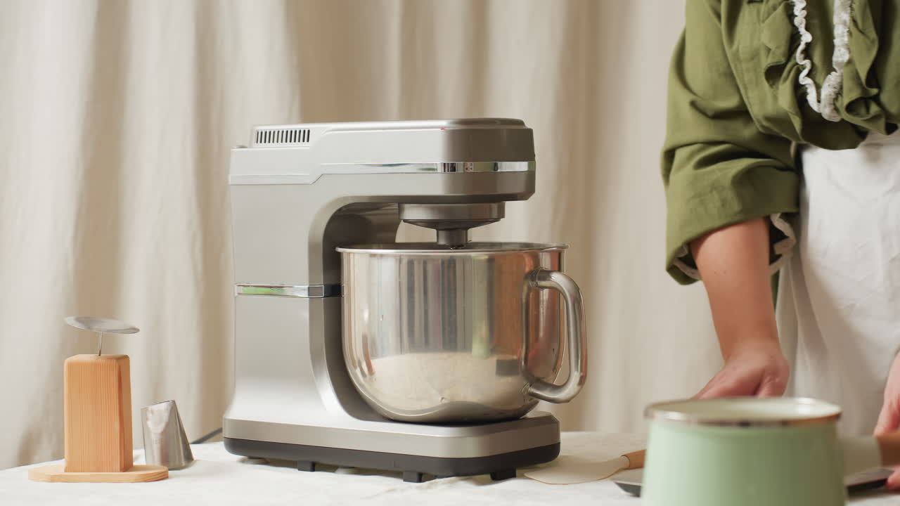 Cook in green top and white apron places mixing bowl into electric stand mixer and removes scale from countertop during baking preparation process in cozy kitchen setting with natural light
