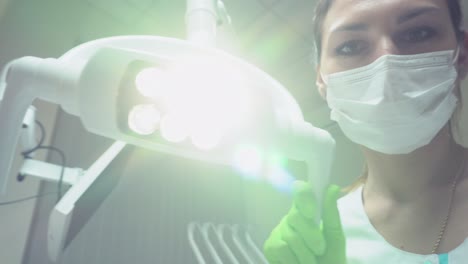 Young-Female-Dentist-in-mask-and-protective-gloves-turns-the-light-of-dental-lamp-on,-standing-upon-a-patient.-Looking-at-camera,-Shot-in-4k