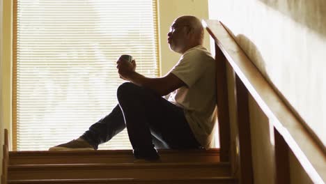 Thoughtful-african-american-senior-man-sitting-at-the-top-of-staircase-in-the-sun-drinking-coffee