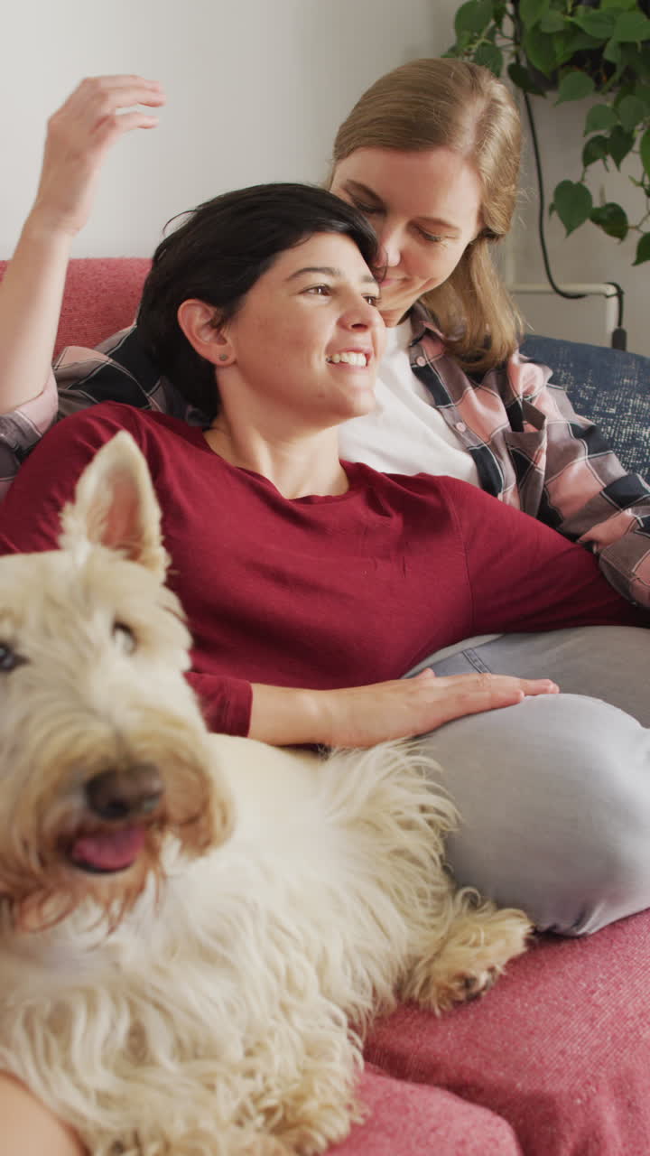 video vertical de una feliz pareja de lesbianas caucásicas abrazándose con un perro en casa