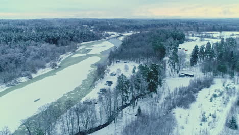 Toma-Panorámica-De-Drones-Aéreos-Sobre-Cabañas-Al-Lado-De-Un-Río-Congelado-Y-Un-Bosque-A-Lo-Largo-Del-Paisaje-Invernal-En-Un-Día-Nublado