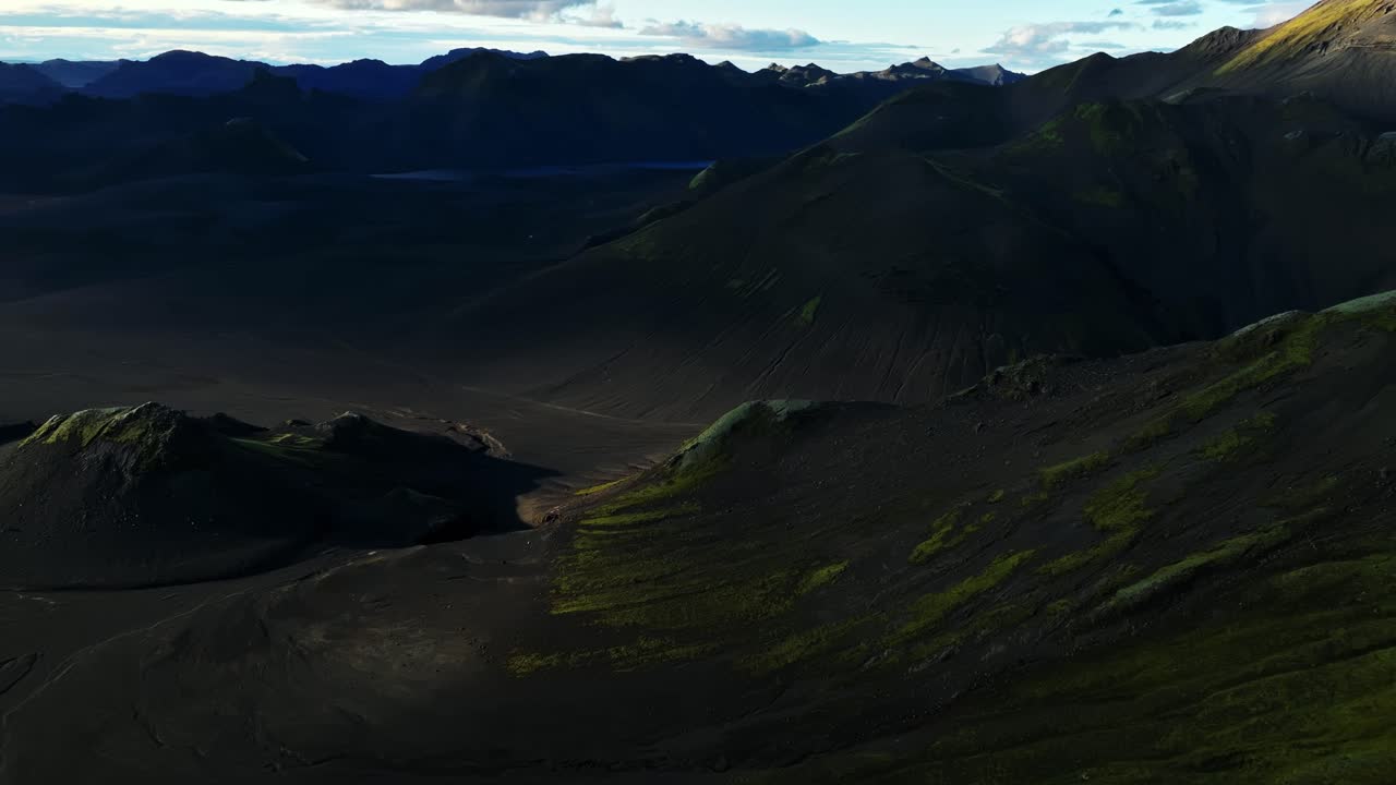 Late-day light reveals dark volcanic slopes and mossy ridges in Iceland’s highlands, casting sweeping shadows across a barren valley surrounded by dramatic mountain forms