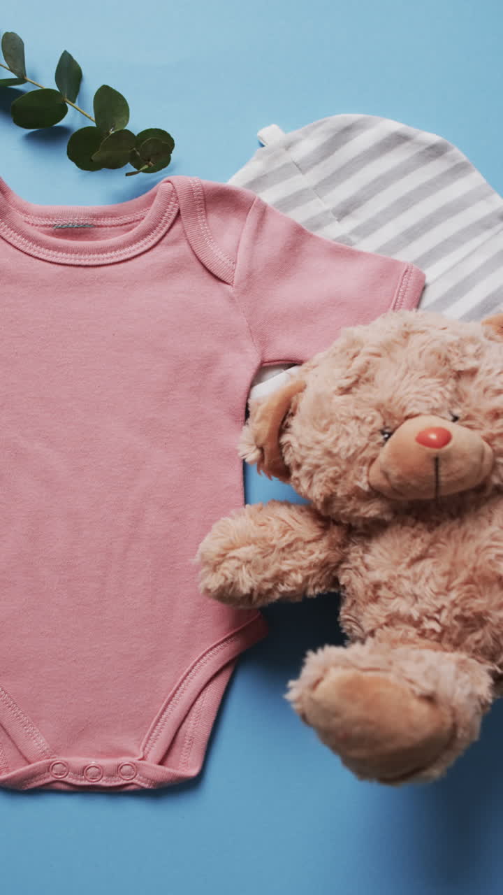 Vertical video of pink baby grow, hat and teddy bear with copy space on blue background