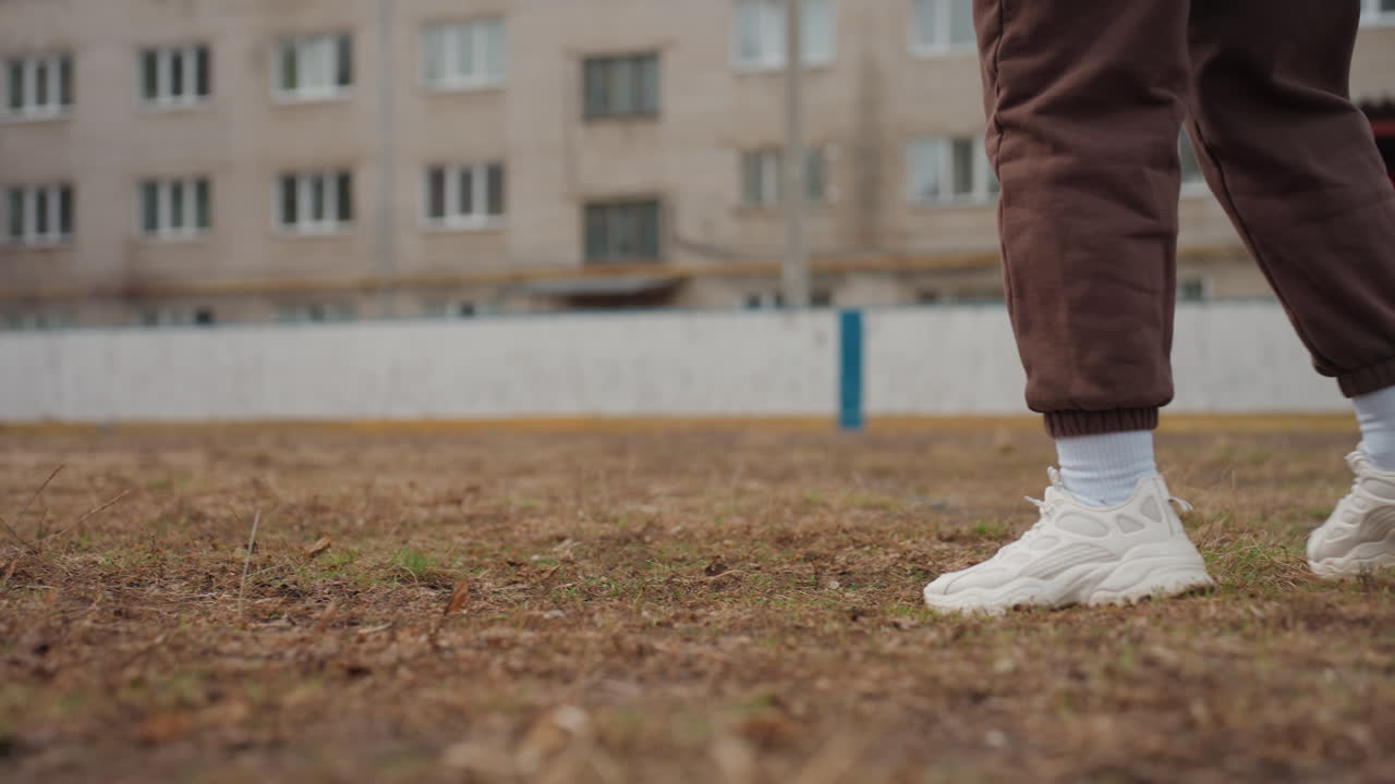 Pies y zapatillas controlando un balón de fútbol sobre hierba húmeda; jugador blanco con ropa de calle moderna que practica toques precisos cerca de pistas de apartamentos, estética urbana minimalista con movimiento enfocado