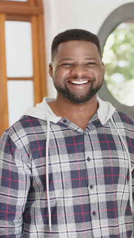 vertical video of happy african american man with beard standing and smiling in sunny house