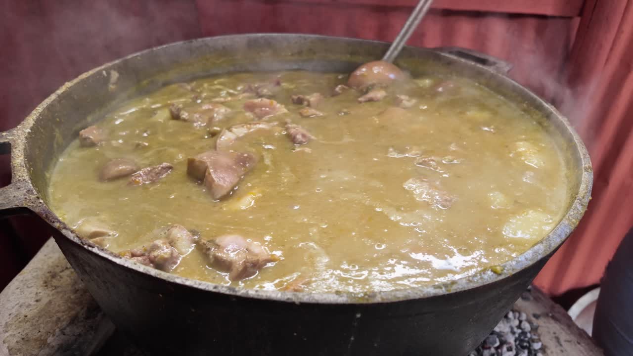 Close-up of a steaming pot cooking Dominican Sancocho in a traditional countryside setting. A metal spoon stirs the meats and vegetables, with steam capturing the rustic essence of this classic dish.