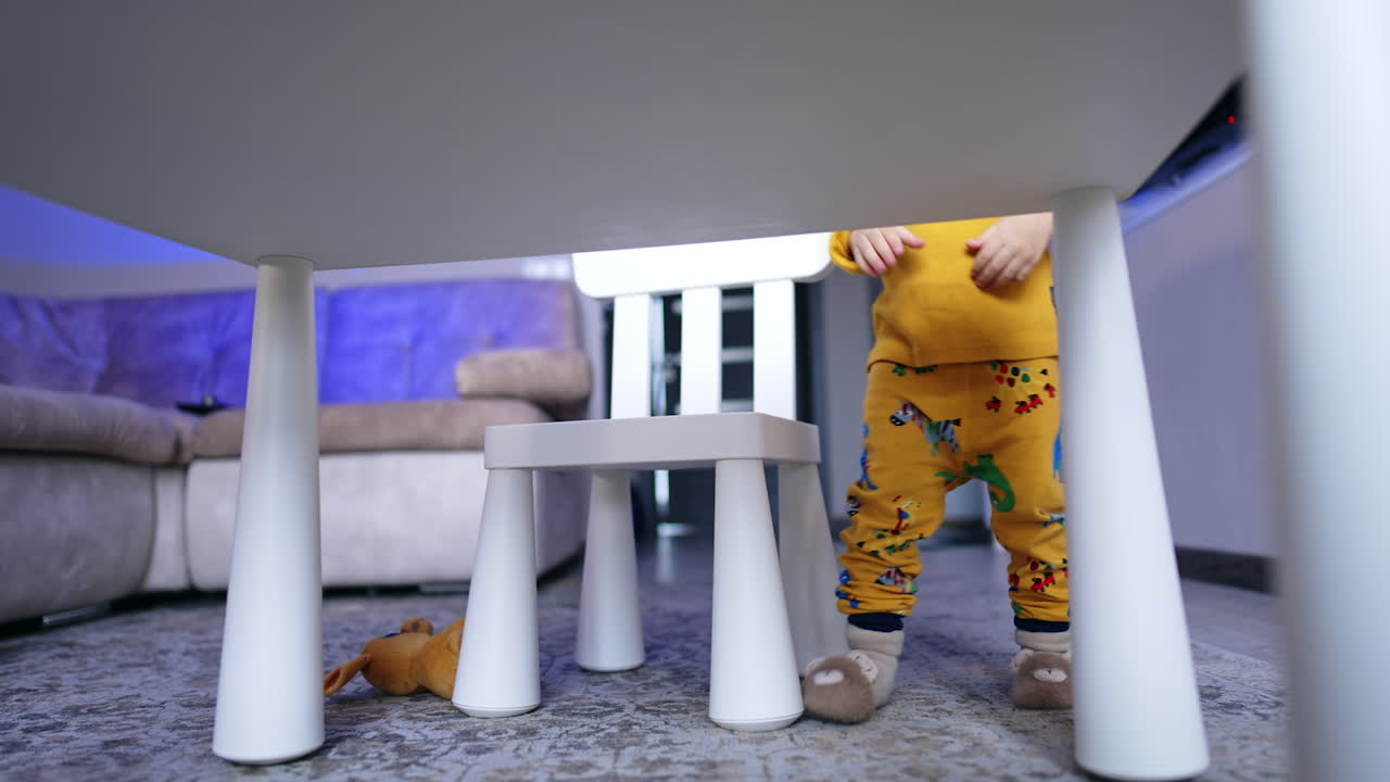 Legs of a baby wearing yellow pants with animals. Baby boy squats to get the dropped puzzle from the desk.