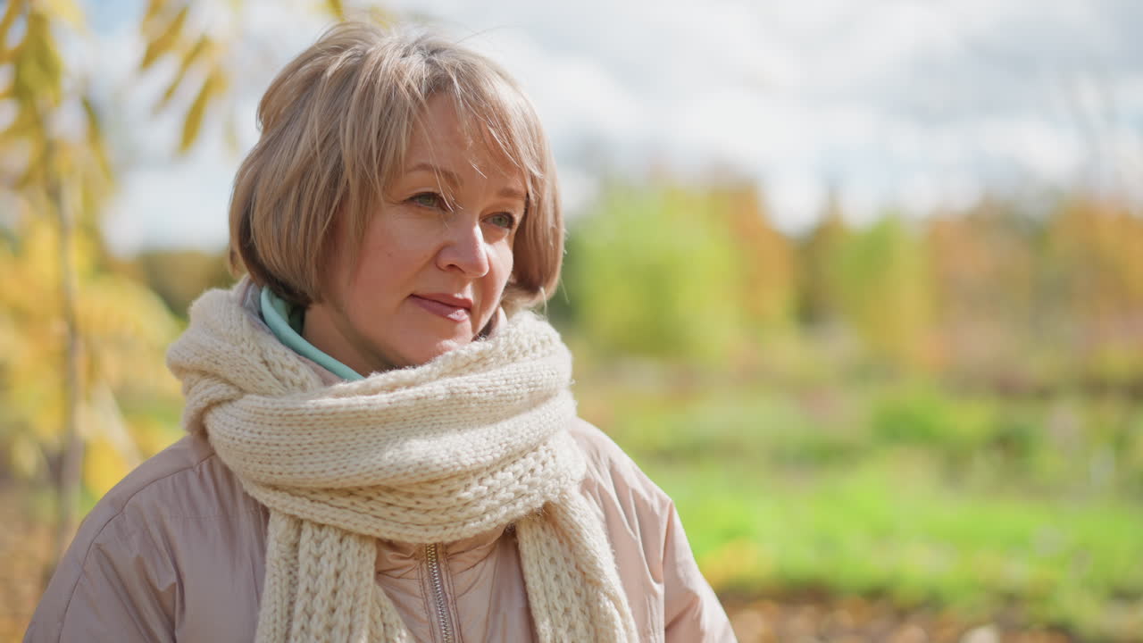 woman in light jacket and cozy scarf standing outdoors holding vibrant yellow autumn leaf with gentle expression while admiring its texture under bright daylight with blurred colorful park background