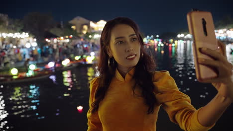 attractive vietnamese woman in traditional yellow dress takes night selfie photo next to river in hoi an, vietnam