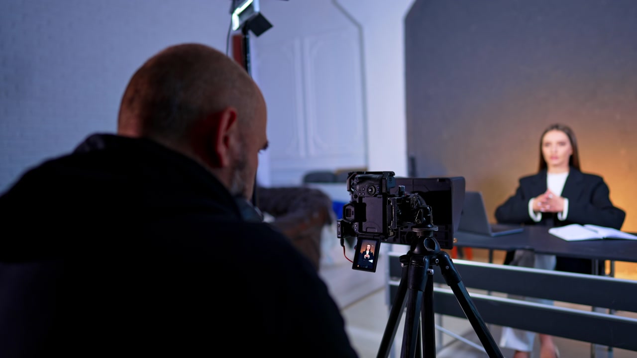 Videografo che regola le impostazioni della fotocamera sul treppiede per un'intervista in studio. Vista da dietro le spalle dell'operatore di ripresa che prepara l'attrezzatura per una presentatrice seduta alla scrivania.