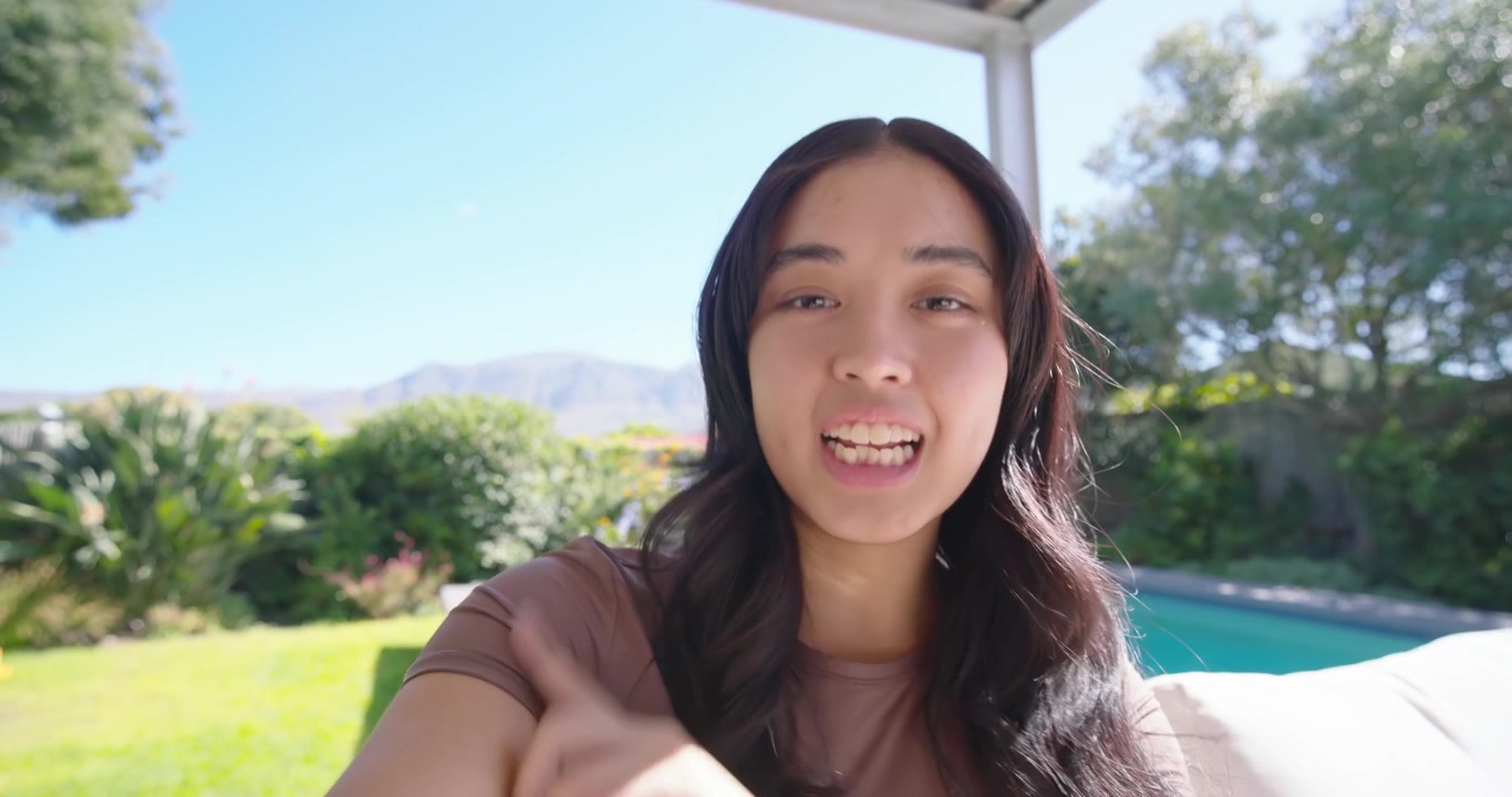 茶色のトップスを着たアジア人成人女性が、スマートフォンで自撮りを始め、プールの方を指差しながら庭について説明している。