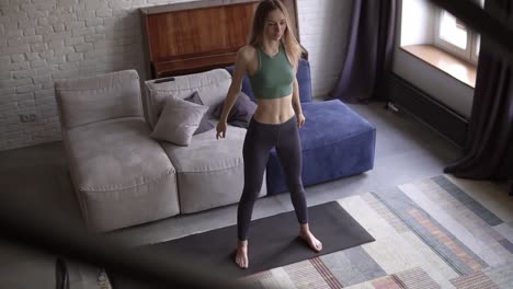 tall woman doing squat exercise on yoga mat in living room at home with couch on the background