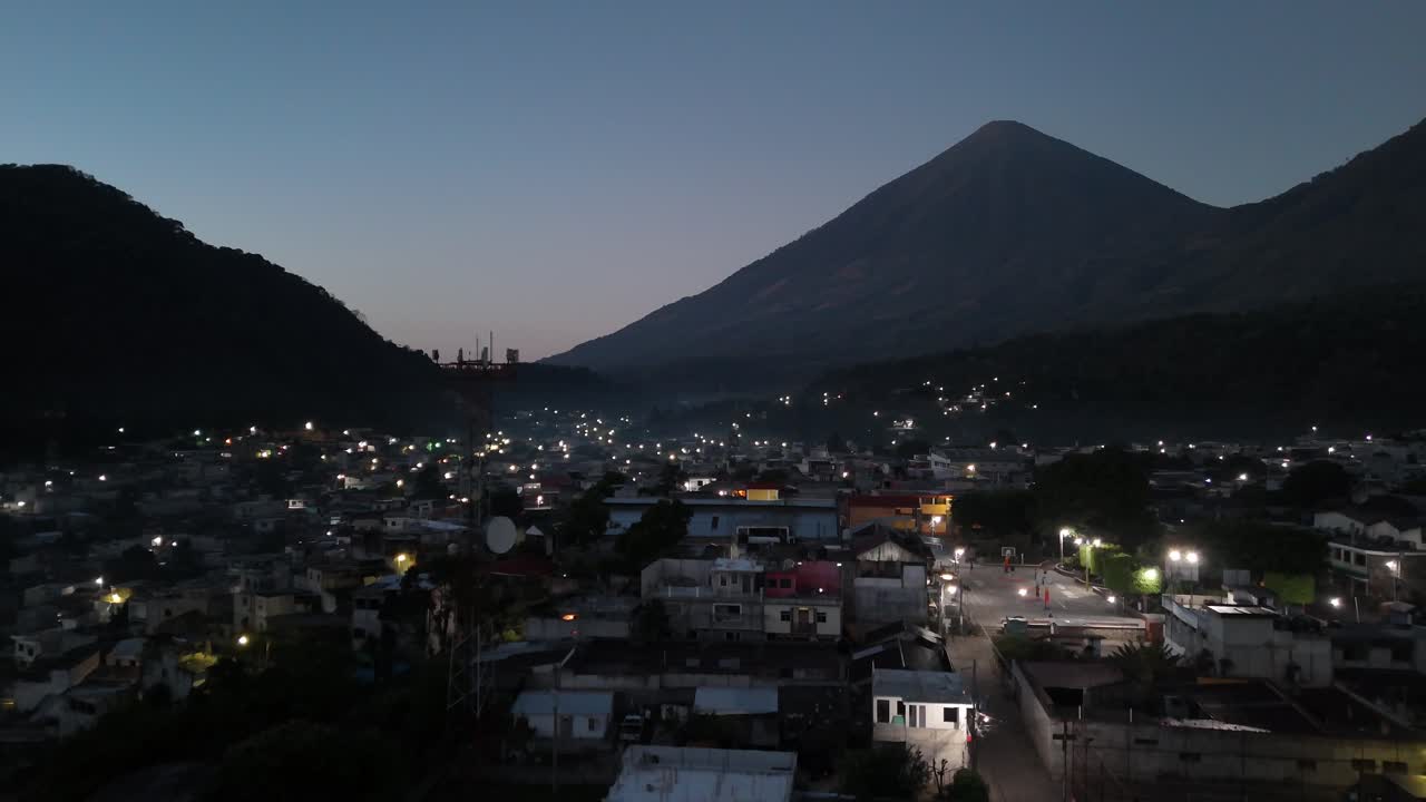 Drone-ilmakuva San Lucas Guatemalasta yöllä esittelee trooppisia asuinalueita, palmuja, värikkään auringonlaskun ja ympäröivät tuliperäiset vuoret, luoden upean Keski-Amerikan maiseman.