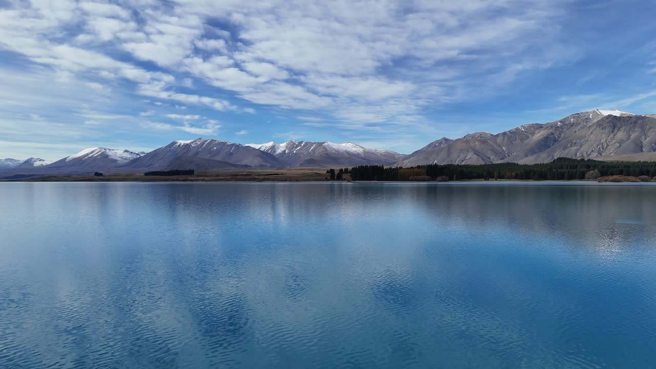 Drone captures Lake Tekapo's calm waters and surrounding mountains under a bright sky, showcasing natural beauty and tranquility