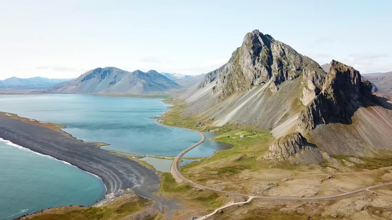 A stunning view of the rugged mountain and serene coastline of Hvalnes, Iceland. The landscape features dramatic cliffs, calm blue waters, and a winding road that blends into the natural beauty