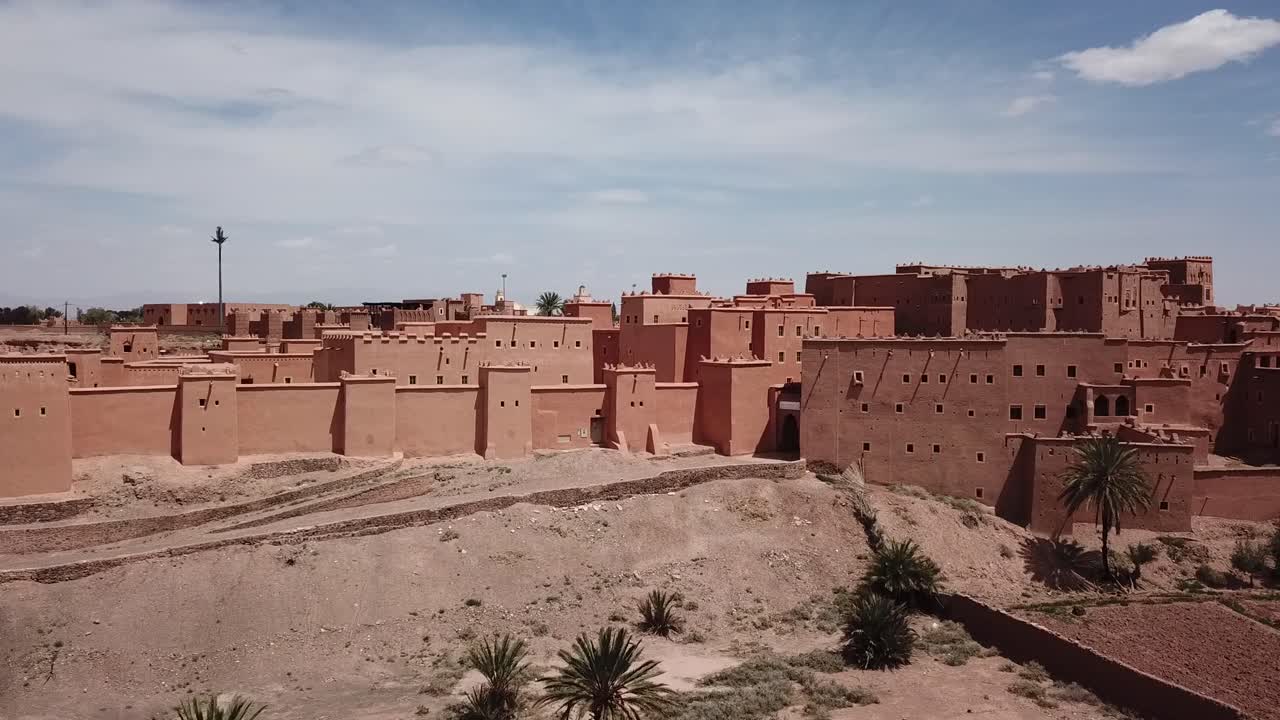 Ampia veduta delle mura di una kasbah marocchina a Ouarzazate, con un primo piano di deserto arido e palme. Forte contrasto tra l'architettura fortificata e il terreno arido, ideale per immagini di viaggio e patrimonio culturale.