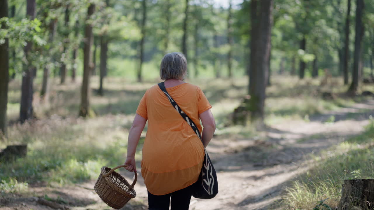 mujeres mayores con camiseta naranja caminando por el bosque verde sosteniendo una canasta de madera y una bolsa negra