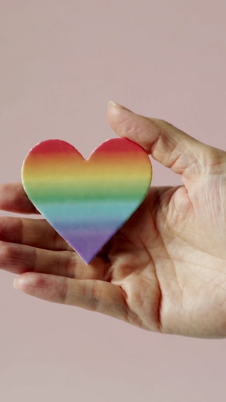 Vertical video: Woman's hand holding rainbow heart on pink backdrop making small shifts for health