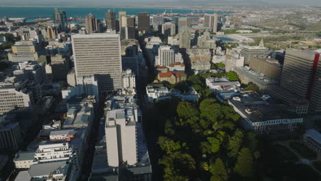 Imágenes-Aéreas-De-Edificios-Alrededor-Del-Jardín-De-La-Empresa-En-El-Centro-De-La-Ciudad.-Superficie-De-Agua-De-Mar-Azul-En-La-Bahía-De-La-Mesa-En-El-Fondo.-Ciudad-Del-Cabo,-Sudáfrica