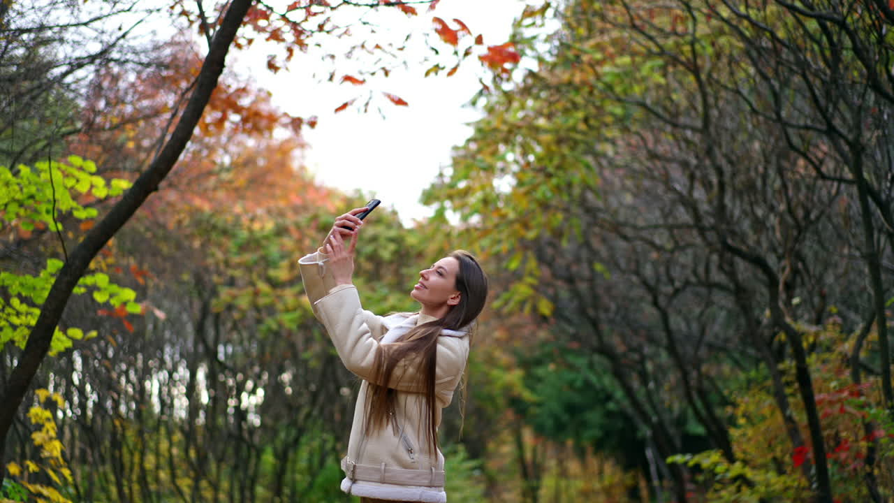 Young attractive long-haired lady taking picture of nature on her phone. Smiling positive woman enjoying beautiful colorful leaves in autumn.