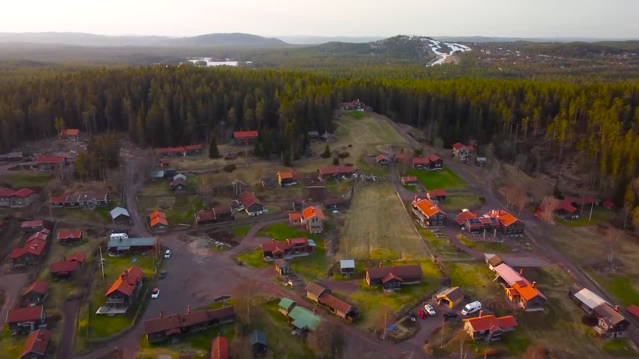 Aerial drone footage of small housing area or a village in Sweden nature during sunset or sunrise. Village with red rooftops is surrounded by dense and thick colorful green pine forest, hills visible.