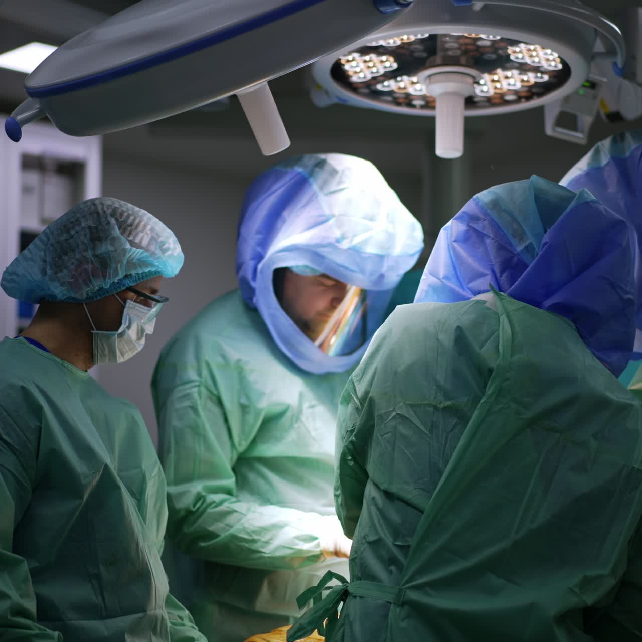 Group of surgeons in protective suits and helmets work under the round lamps in surgery room. Team of doctors cooperate at work