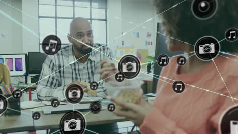 Animation-of-connected-icons-over-diverse-coworkers-discussing-while-eating-donuts-at-desk