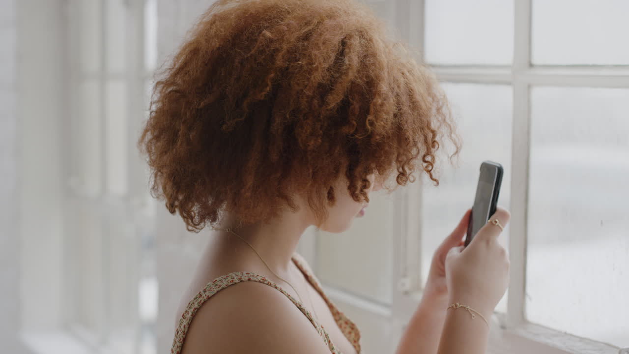 retrato de una joven alegre tomando una foto usando tecnología de cámara móvil de teléfono inteligente pegajosa de raza mixta estilo de cabello afro femenino de pie junto a la ventana