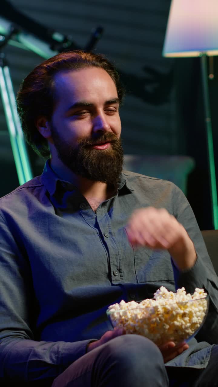 Vertical video Man laughing at funny shows on television, eating snack, sitting on couch