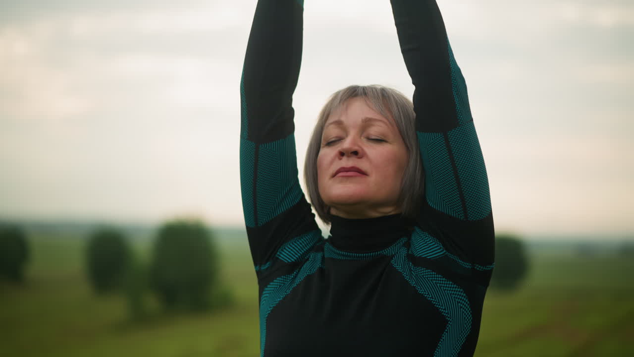 Middle-aged woman with arms raised brings hands together in meditation, practicing mindfulness outdoors in a grassy misty field under cloudy sky in the background