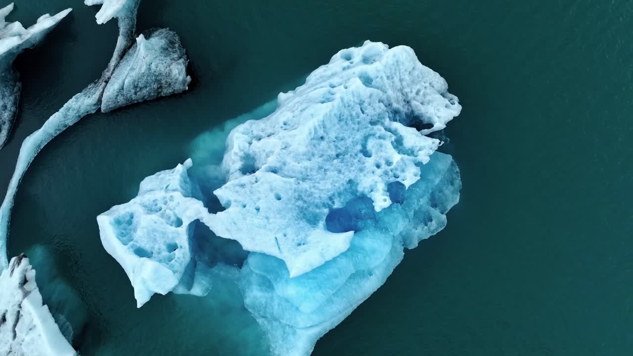 Top-down descending drone shot of a sculpted blue iceberg drifting in dark glacial water, revealing textured ice layers, deep pockets, and crisp Arctic tones in a calm frozen landscape