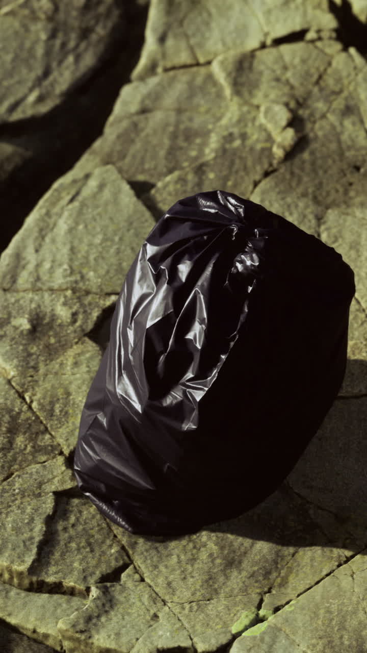 Black trash bag rests on rocky shoreline under soft sunlight