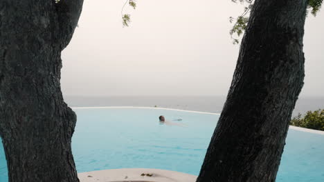 Wide-push-in-shot-of-young-woman-swimming-in-infinity-pool,-overlooking-vast-ocean