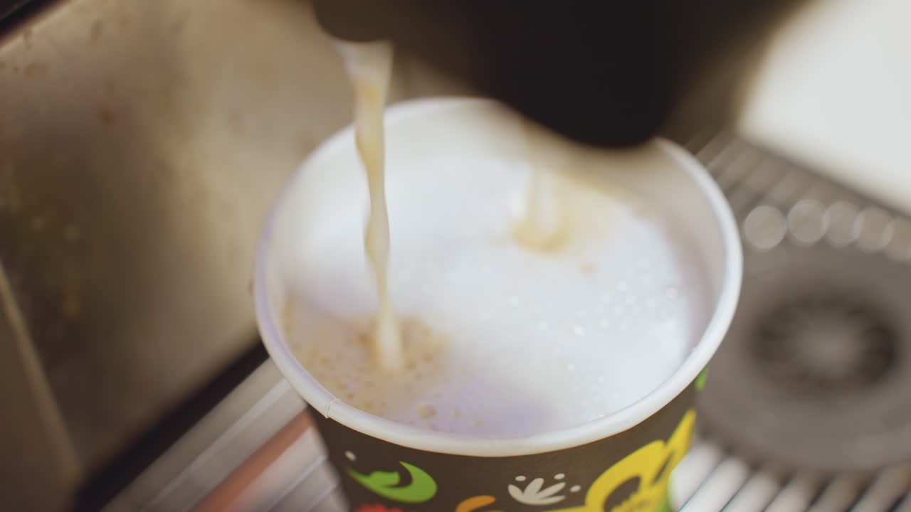 Primer plano de una taza de café recibiendo un vertido de café con leche, un chorro de espresso que se une a la espuma de leche al vapor en un colorido vaso de papel en el concurrido mostrador de una cafetería, con la boquilla de la máquina de acero inoxidable visible, una sugerencia de aroma cálido y el servicio matutino.