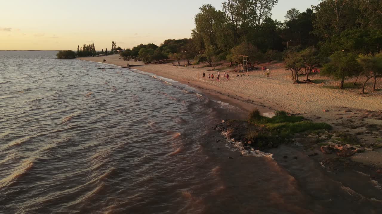 vuelo aéreo hacia atrás de la costa de fray bentos con un grupo de personas jugando voleibol durante la puesta de sol dorada en uruguay - disfrutando de vacaciones y vacaciones en verano