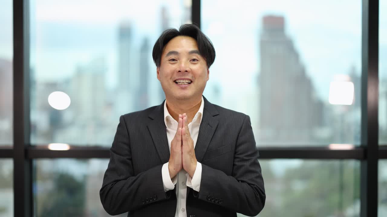 Confident Asian businessman bows with wai gesture, smiling in bright, contemporary office setting