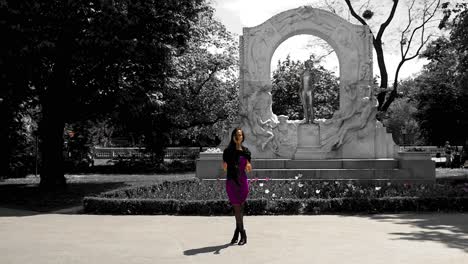 una chica asiática morena juega con el cabello de pie frente al monumento a johann strauss en viena, sonriendo.